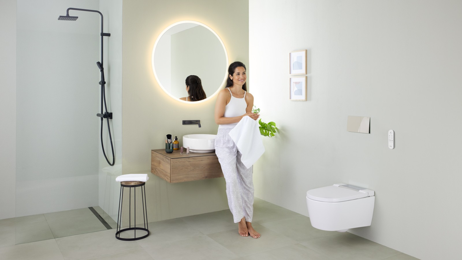 Femme appuyée sur un lavabo dans une salle de bains avec douche Geberit AquaClean Sela, lavabo Geberit VariForm et meubles. Femme appuyée sur un lavabo dans une salle de bains avec douche Geberit AquaClean Sela, lavabo Geberit VariForm et meubles.