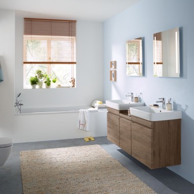 Salle de bains familiale aux murs bleu clair avec meubles en noyer carya, armoire de toilette, plaque de déclenchement et céramiques sanitaires de Geberit